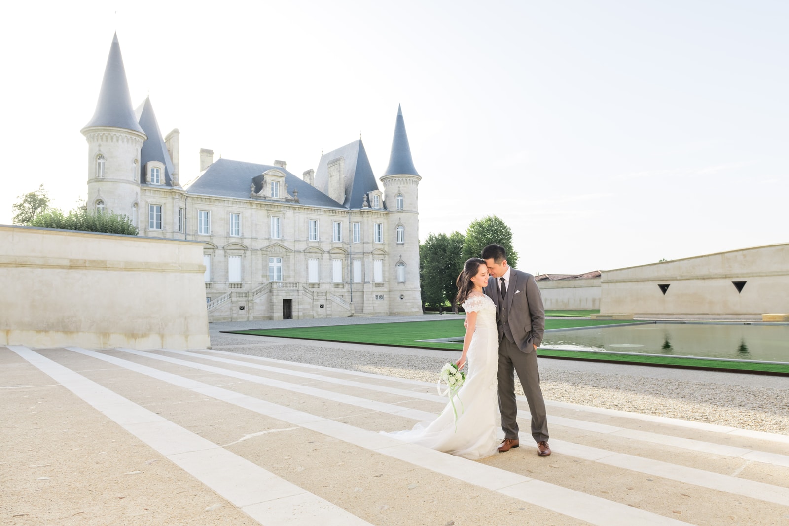 Les mariés, minuscules, avancent sur l'esplanade géométrique devant la façade d'un grand château
