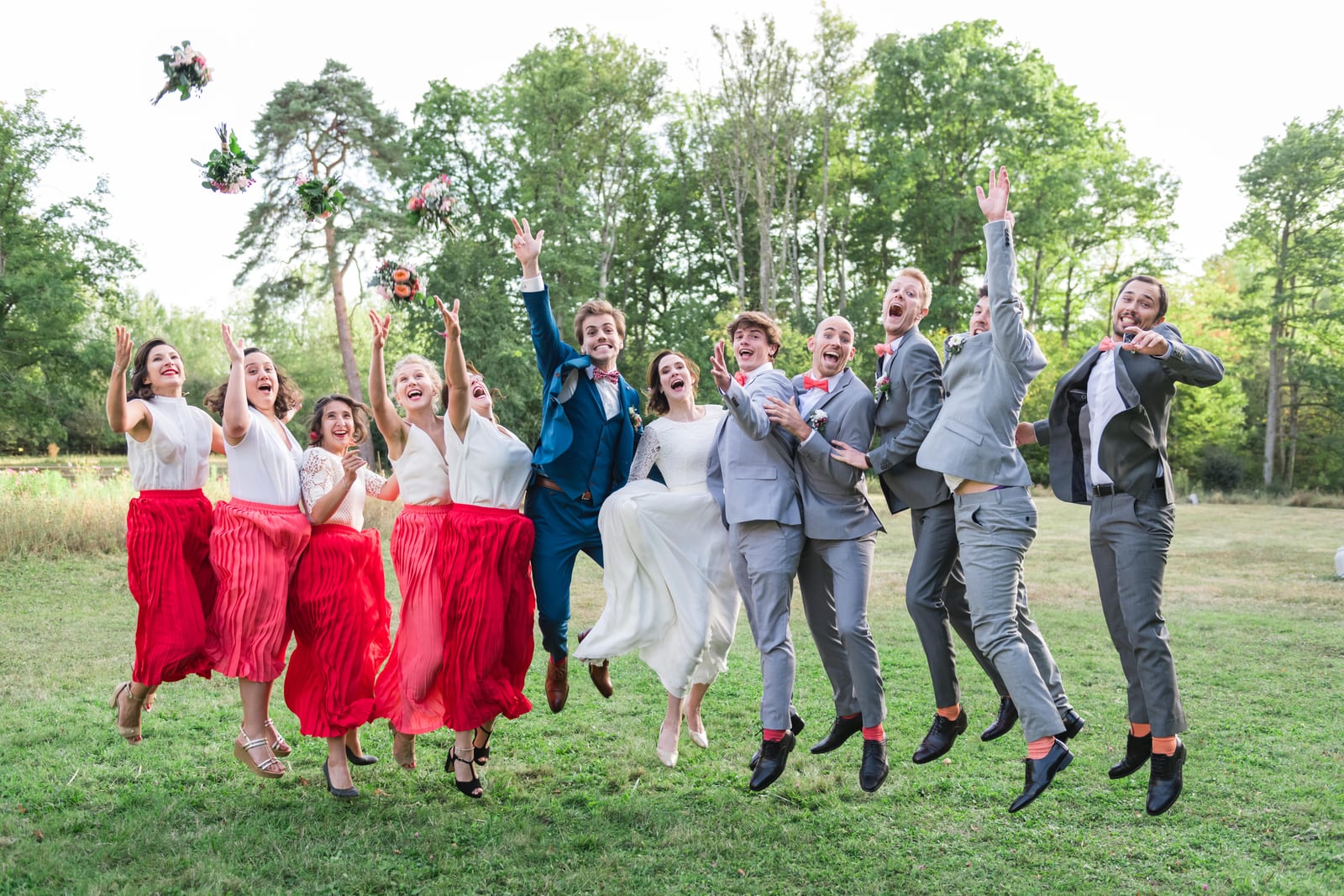 Les mariés et tout leur cortège sautent ensemble en lançant les bouquets, demoiselles d'honneur en jupes rouges