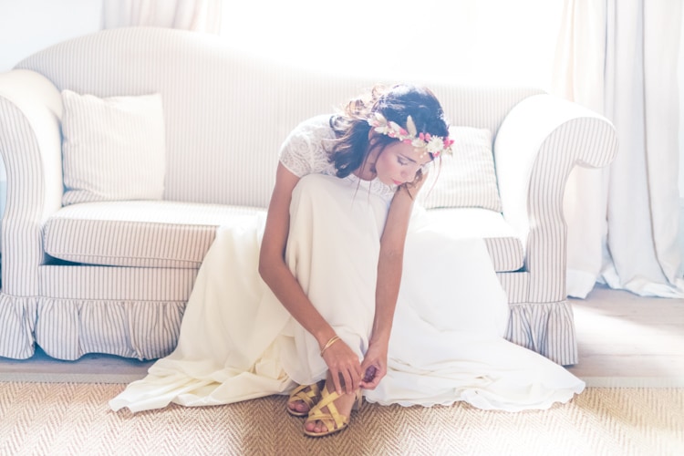 La mariée, couronne de fleurs séchées dans les cheveux, attache ses sandales dorées assise devant un canapé rayé