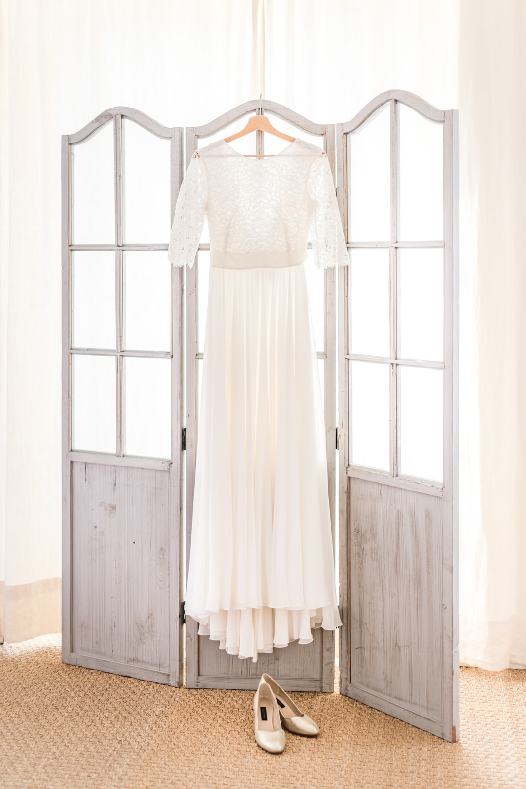 La robe de mariée suspendue à un paravent de bois clair, escarpins posés au sol dans la lumière du matin
