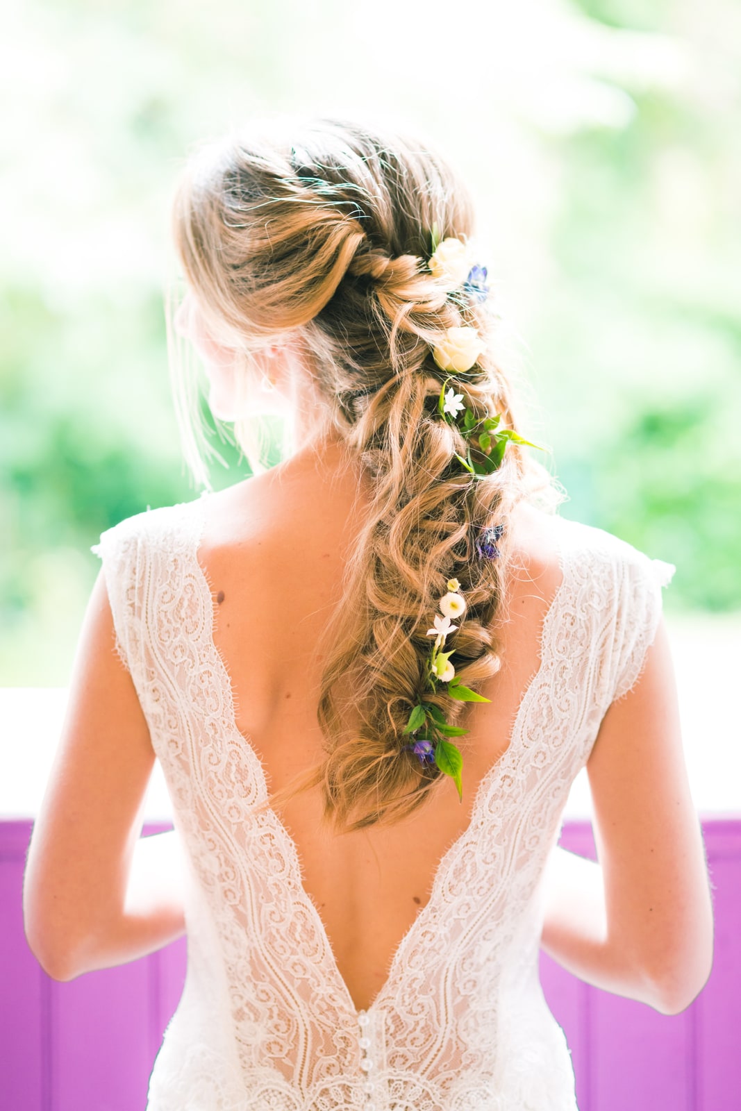 De dos, la mariée en robe de dentelle au dos ouvert, longue tresse parsemée de fleurs fraîches
