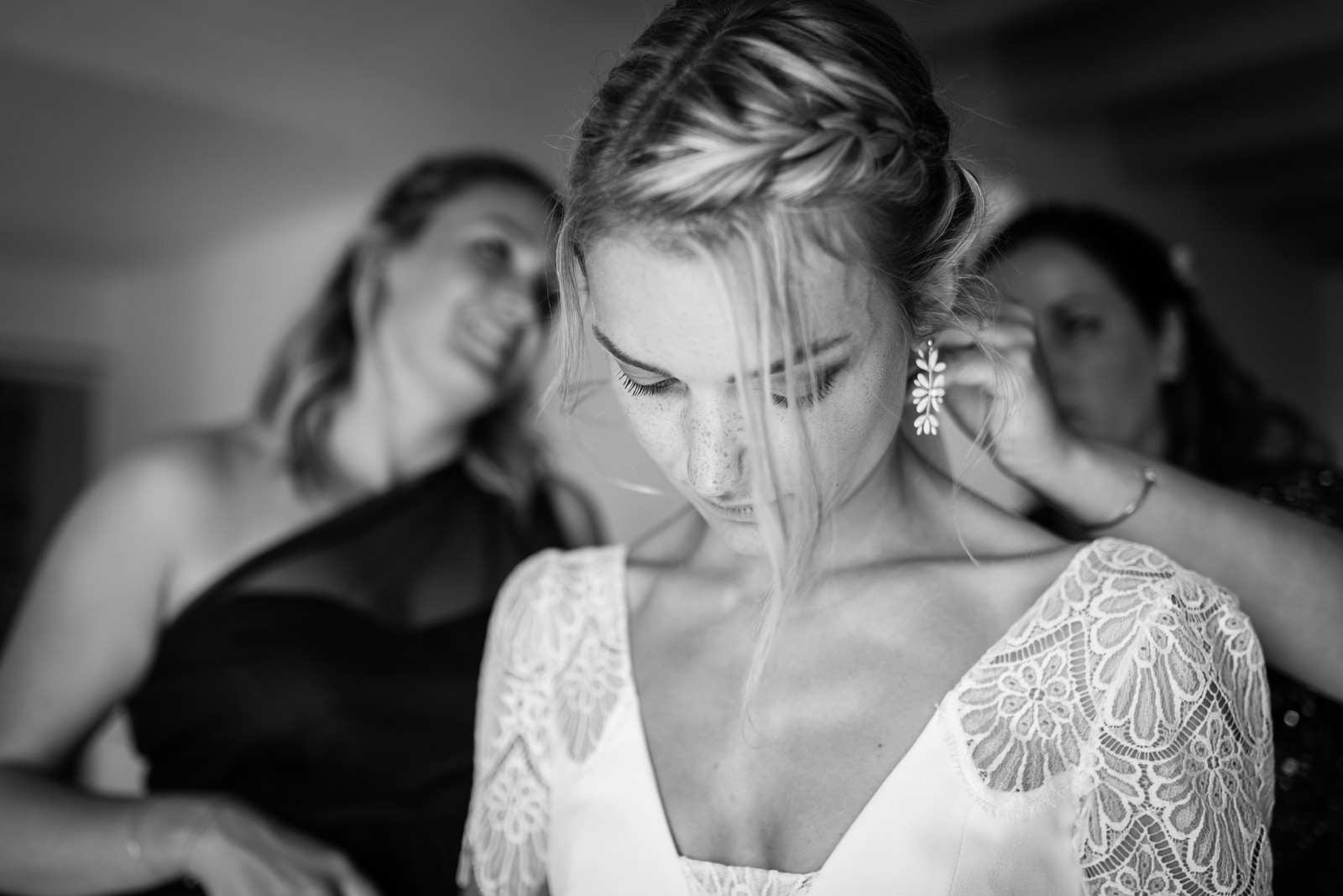 Une proche agrafe la boucle d'oreille de la mariée en robe de dentelle, sourires complices autour d'elle