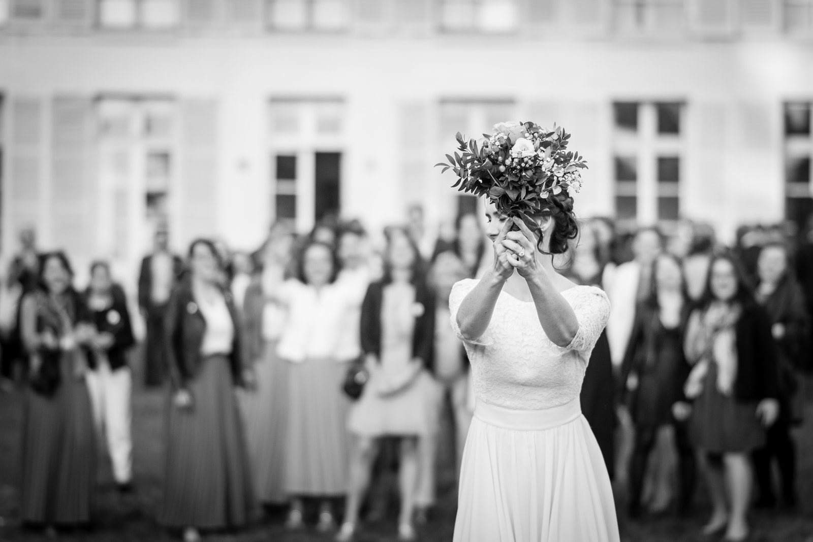 La mariée cache son visage derrière son bouquet, face à la haie formée par les invités devant la façade du château