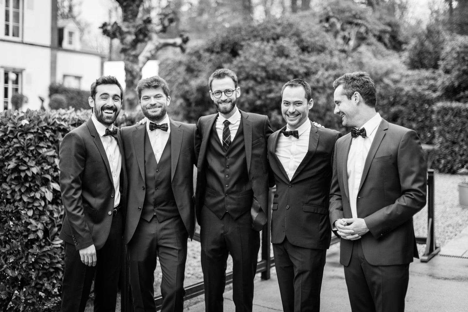 Cinq hommes en smoking et nœud papillon posent en riant devant une haie taillée